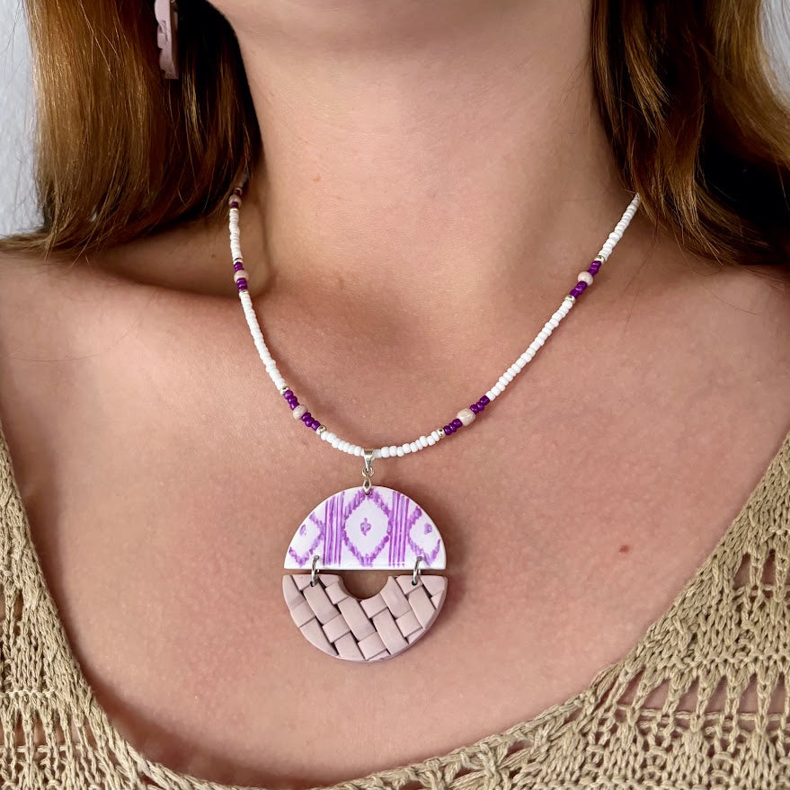 Modelo luciendo collar artesanal Arrels Alba con colgante de arcilla polimérica, estampado de llengos en lila y textura de llata en beige, cadena de cuentas de vidrio, colección Esencia Mallorquina
