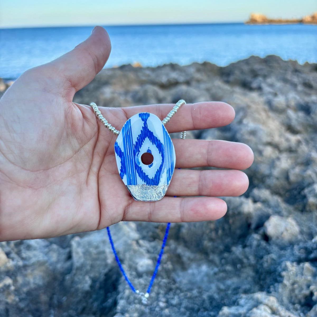 Primer plano de un colgante artesanal de la colección Esencia Mallorquina con diseño de lenguas azules y blancas, mostrado en la palma de la mano para apreciar su tamaño y la textura del cordón de cuentas plateadas y azules, con el mar de fondo.