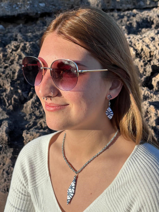 Modelo luciendo los pendientes cortos artesanales Llengos Sal - Rombo corto de arcilla polimérica en azul y blanco con fornitura bañada en plata y collar de cuentas plateadas con colgante rombo, con gafas de sol y fondo de roca natural de Mallorca. Colección Esencia Mallorquina de Pm_Art.