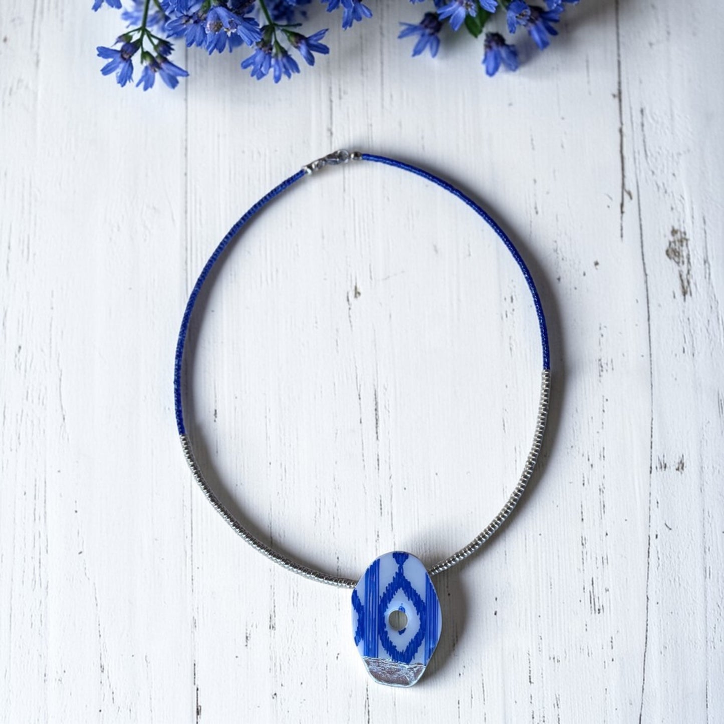 Gargantilla artesanal de cuentas azules y plata, con colgante de arcilla polimerica con estampado de lenguas mallorquinas pintado a mano en azul y blanco sobre fondo de madera clara con flores azules.