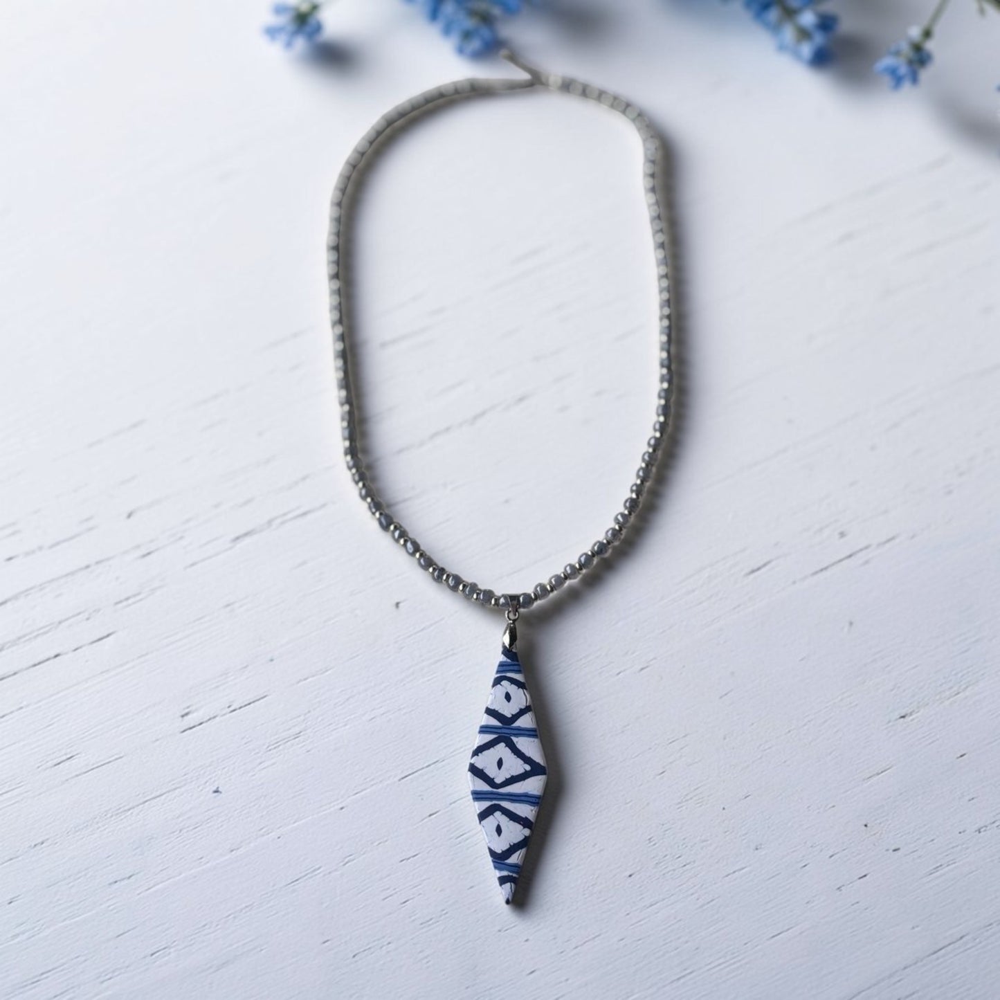 Collar artesanal Llengos Sal con colgante rombo alargado de arcilla polimérica en azul marino y blanco y cuentas de cristal azul claro y plateado con cierre de acero inoxidable, sobre fondo de madera blanca con flores azules. Colección Esencia Mallorquina de Pm_Art.
