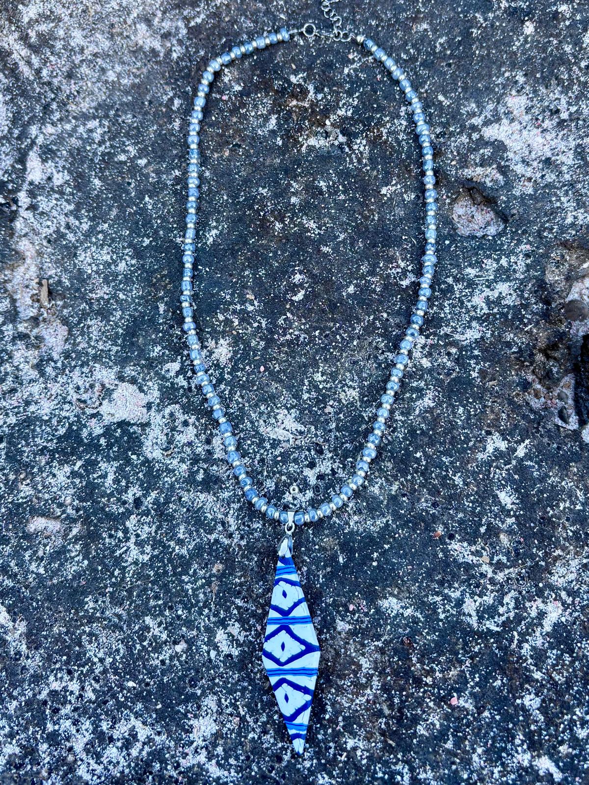 Collar artesanal Llengos Sal con colgante rombo alargado de arcilla polimérica en azul marino y blanco y cuentas de cristal en azul claro y plateado, sobre roca oscura de Mallorca al sol. Colección Esencia Mallorquina de Pm_Art.
