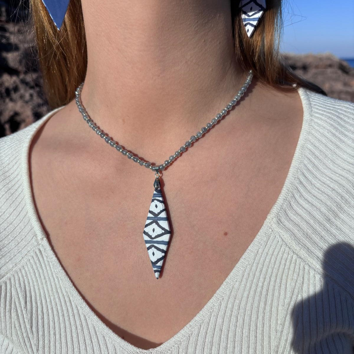 Modelo luciendo el collar artesanal Llengos Sal con colgante rombo alargado de arcilla polimérica en azul marino y blanco y cuentas de cristal en azul claro y plateado, combinado con pendientes a juego, jersey blanco y fondo de costa rocosa de Mallorca. Colección Esencia Mallorquina de Pm_Art.
