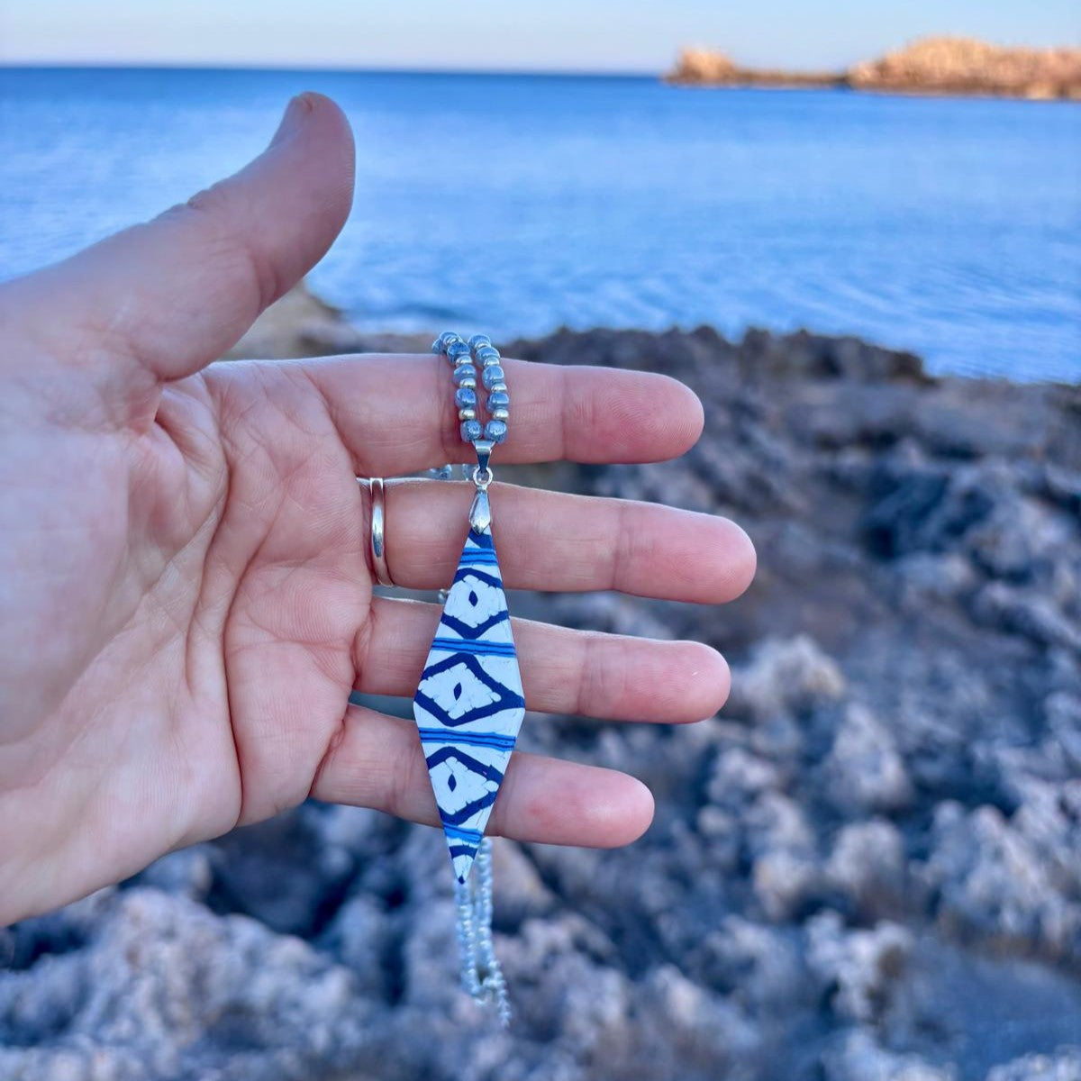 Collar artesanal Llengos Sal con colgante rombo alargado de arcilla polimérica en azul marino y blanco y cuentas de cristal en azul claro y plateado, sostenido en mano con la costa rocosa y el mar Mediterráneo de Mallorca al fondo. Colección Esencia Mallorquina de Pm_Art.
