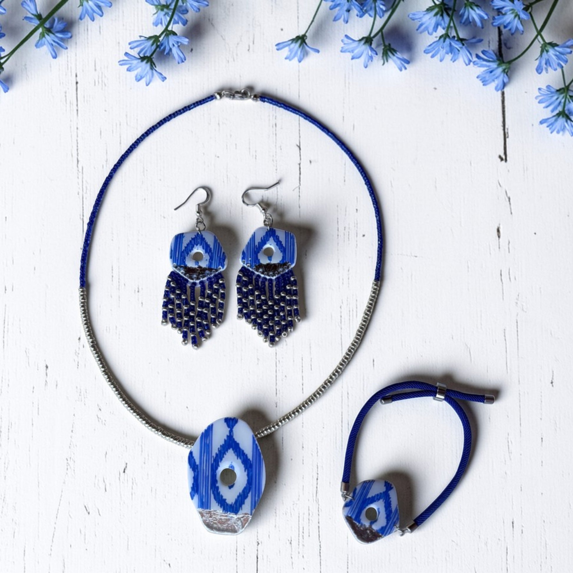 Set de collar, pendientes y pulsera de arcilla polimérica con diseño de lenguas mallorquinas en azul, blanco y plata. Fotografía del producto sobre fondo claro con detalles de flores azules.