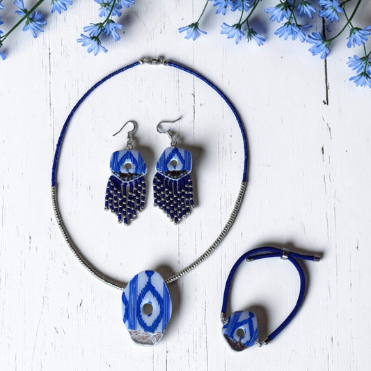 Set de collar, pendientes y pulsera de arcilla polimérica con diseño de lenguas mallorquinas en azul, blanco y plata. Fotografía del producto sobre fondo claro con detalles de flores azules.
