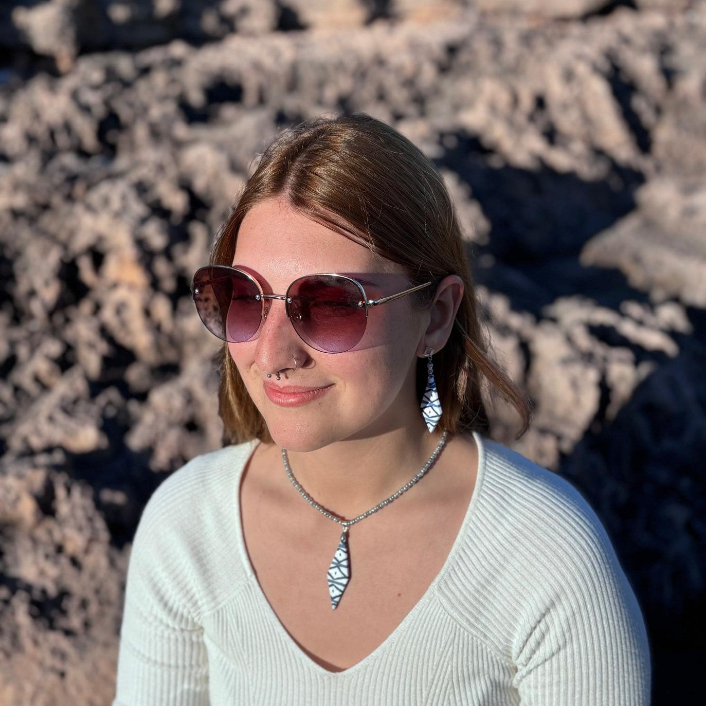 Modelo luciendo el collar artesanal Llengos Sal con colgante rombo alargado de arcilla polimérica en azul marino y blanco y cuentas de cristal en azul claro y plateado, combinado con pendientes a juego, gafas de sol y jersey blanco con fondo de roca natural de Mallorca. Colección Esencia Mallorquina de Pm_Art.
