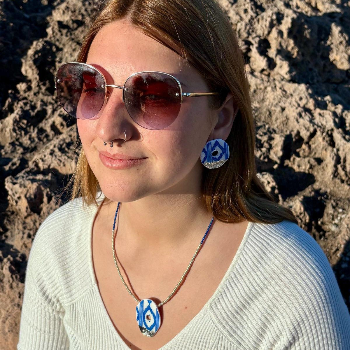 Modelo luciendo el conjunto mediterráneo de colgante y pendientes de botón con estampado de lenguas mallorquinas azules. Fotografía de estilo de vida capturada en un entorno natural de costa que resalta la elegancia artesanal de las piezas.