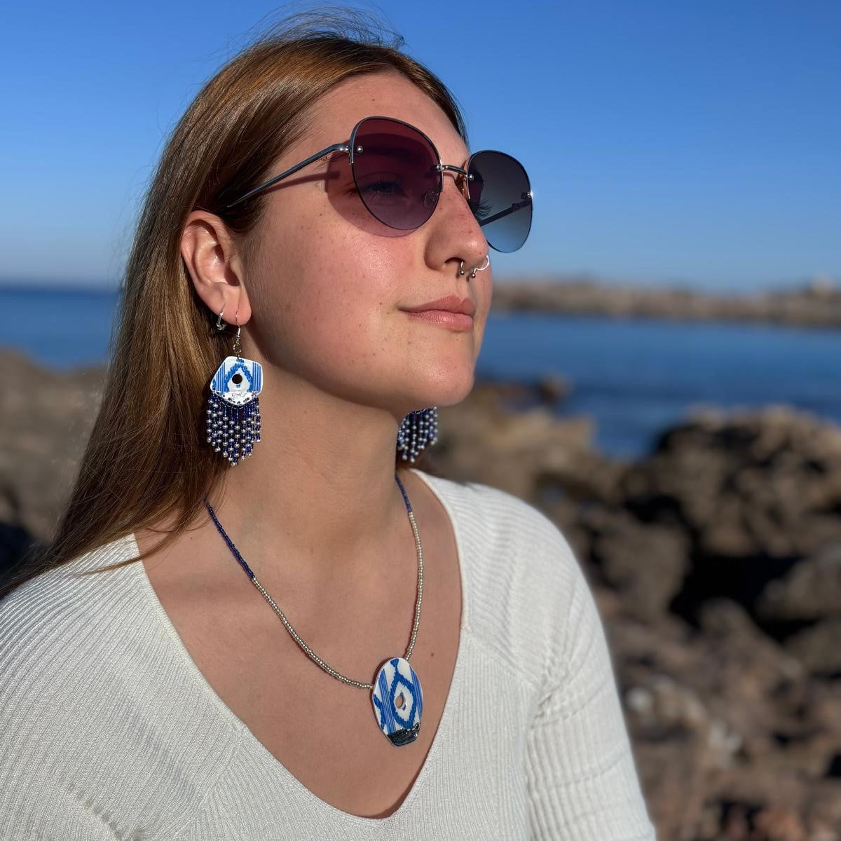 Modelo luciendo pendientes y collar artesanales azul y blanco en la playa. Se aprecia el collar corto y los pendientes largos con flecos de cuentas, resaltando el estilo mediterráneo de la colección Esencia Mallorquina