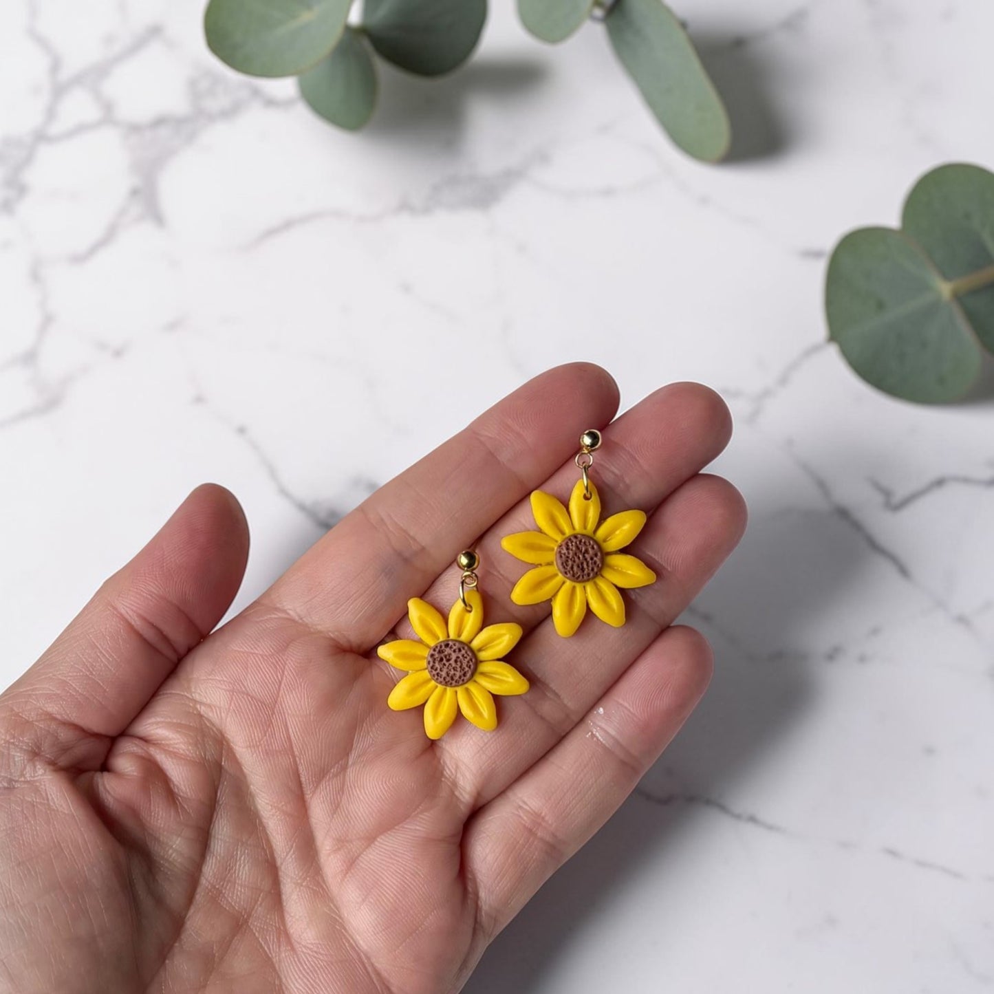 Mano sosteniendo pendientes artesanales de girasol amarillo de arcilla polimérica mostrando tamaño real sobre fondo de mármol con eucalipto