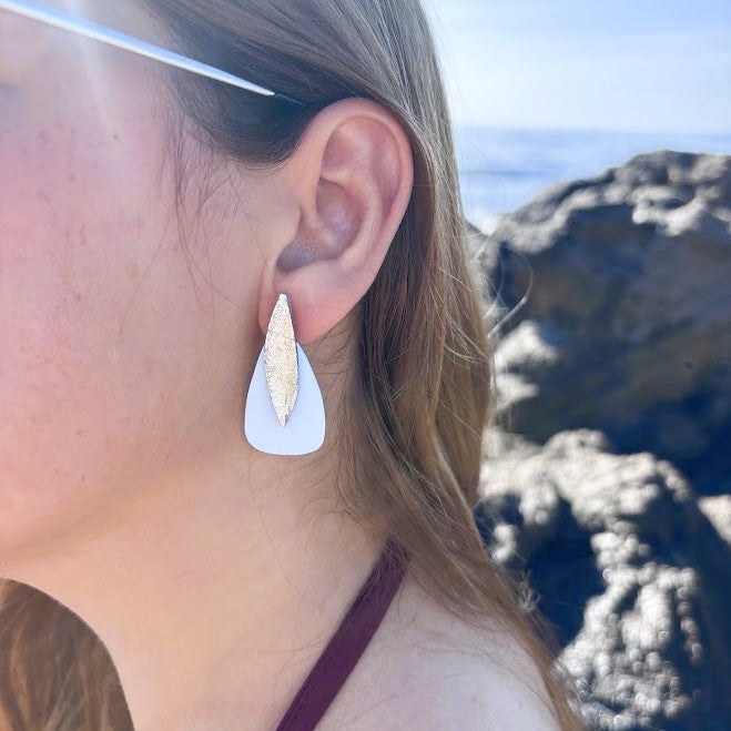Primer plano de los pendientes artesanales en oreja modelo sail en color azul nube y dorado con fondo de rocas y mar
