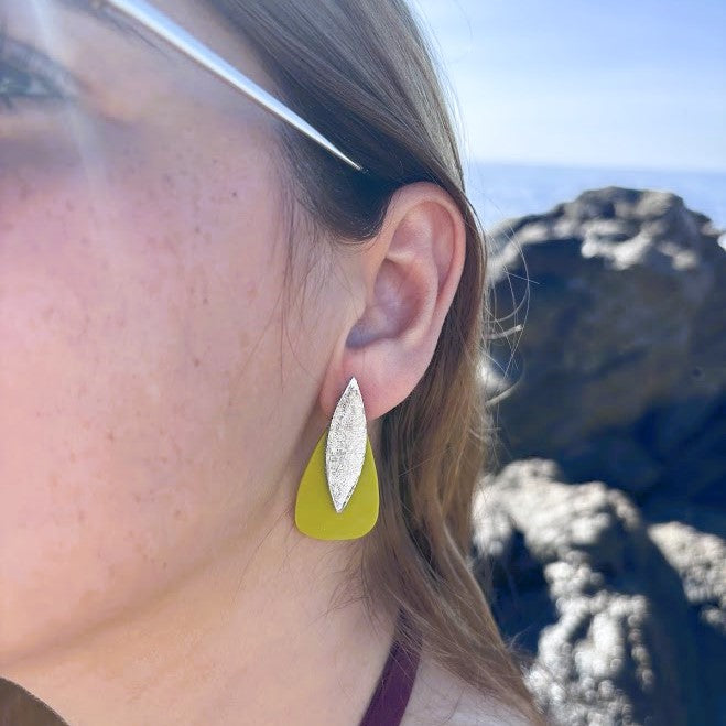 Modelo luciendo pendientes Sail color verde oliva de arcilla polimérica, vista lateral con fondo de rocas y mar