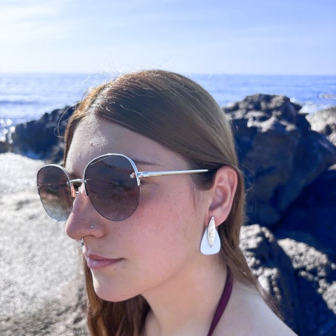 Plano de perfil de modelo luciendo los pendientes artesanales  Sail de color azul nube y plata con fondo de mar y rocas