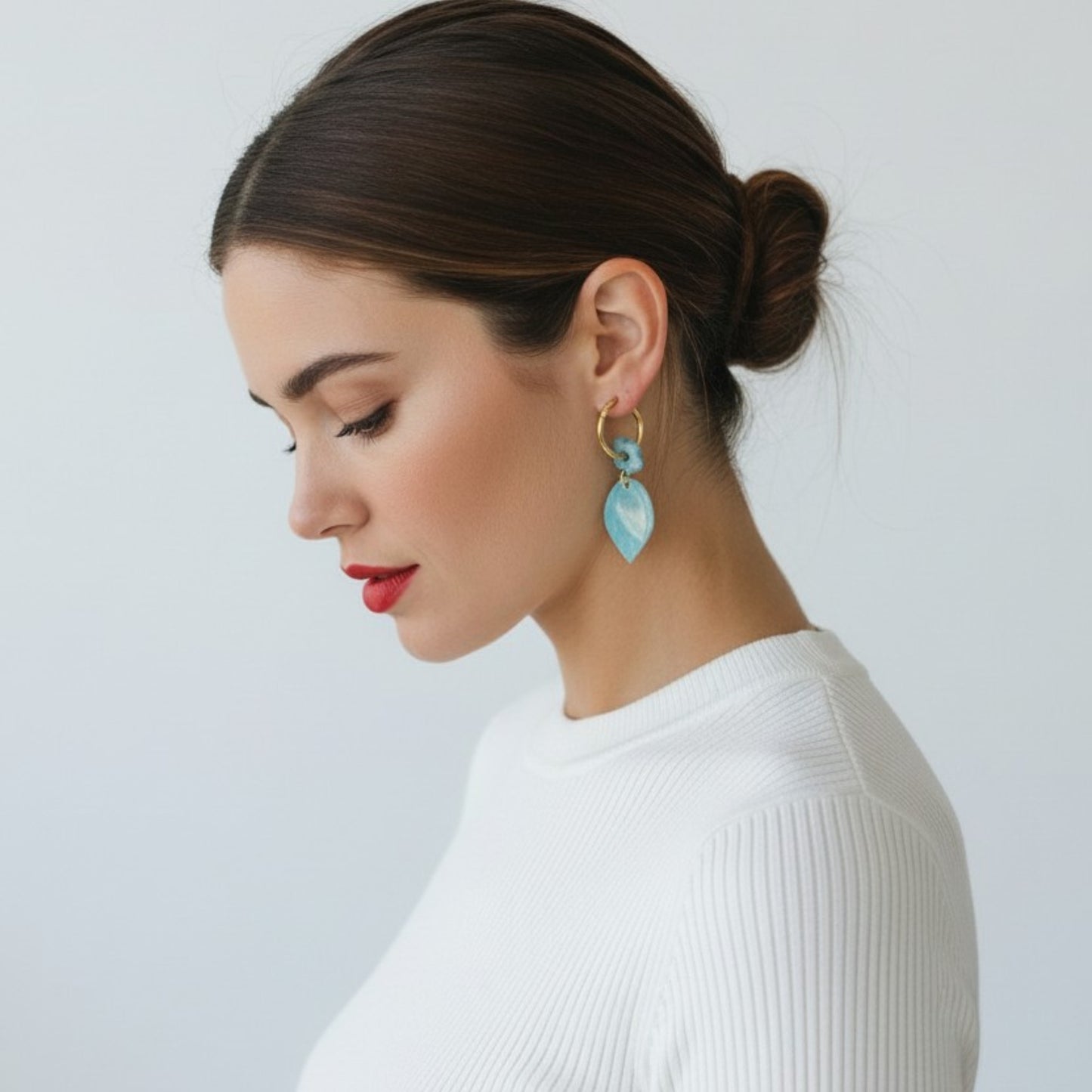Mujer con moño luciendo pendientes artesanales Blue Leaf de arcilla polimérica azul turquesa y oro sobre jersey blanco