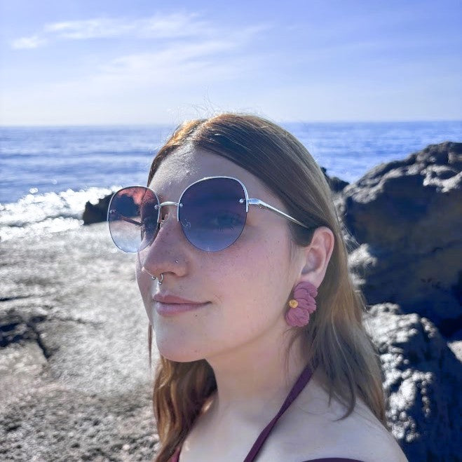 Modelo luciendo pendientes Sollerich de media flor rosa humo oscuro en arcilla polimérica, con el mar de fondo