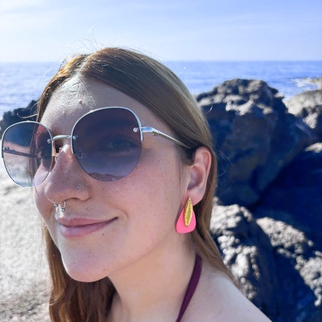 Modelo llevando pendientes Sail color rosa coral de arcilla polimérica, diseño bohemio, con fondo de rocas y mar