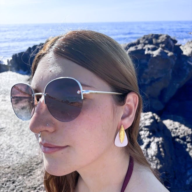 Modelo luciendo pendientes modelo Sail de color beig y dorado con fondo de rocas y mar