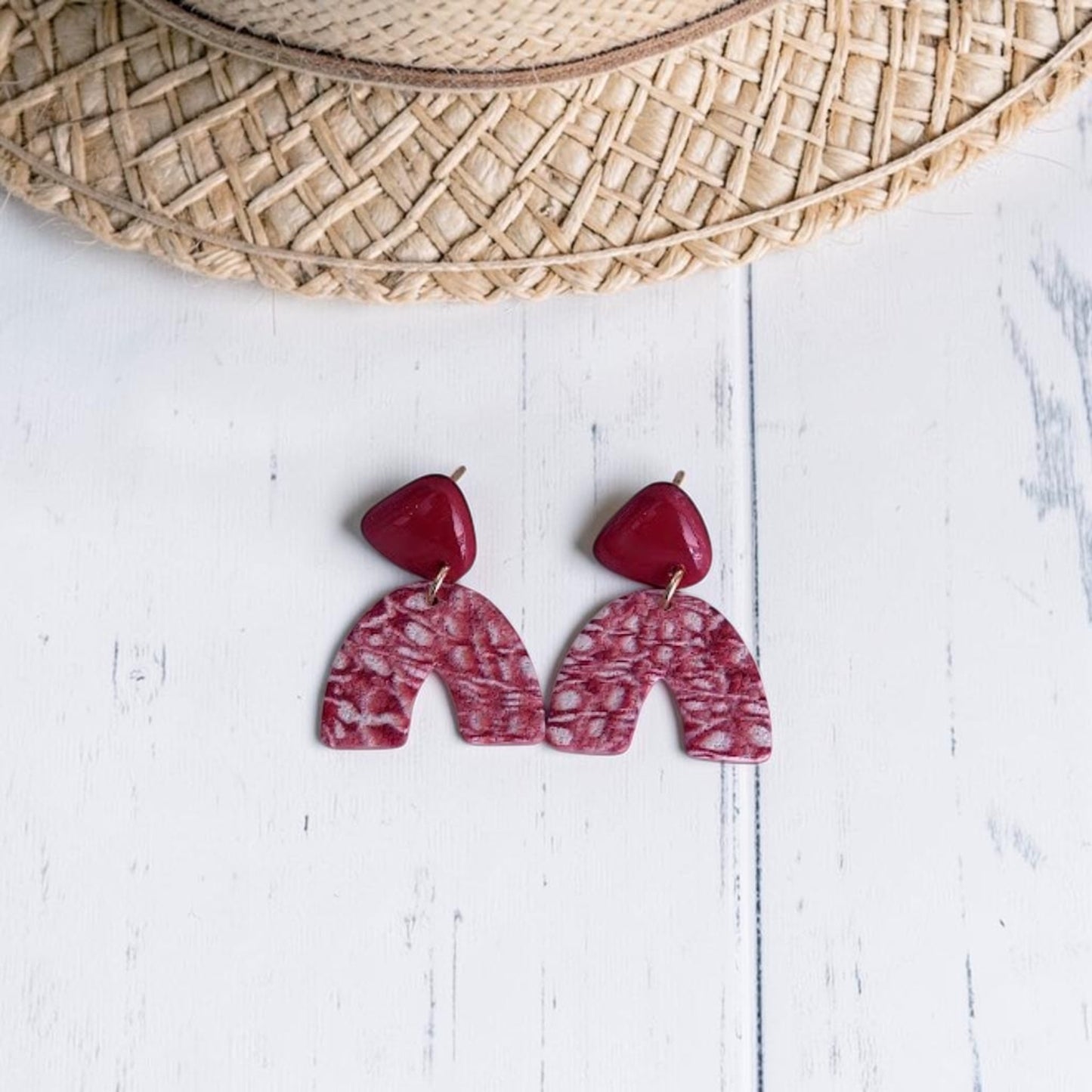 Pendientes artesanales de arcilla polimérica con diseño de arco con estampado marmoleado en color teja y beig y pieza superior color teja acabada con resina UV, presentados sobre fondo de madera blanca con un detalle de un sombrero de rafia