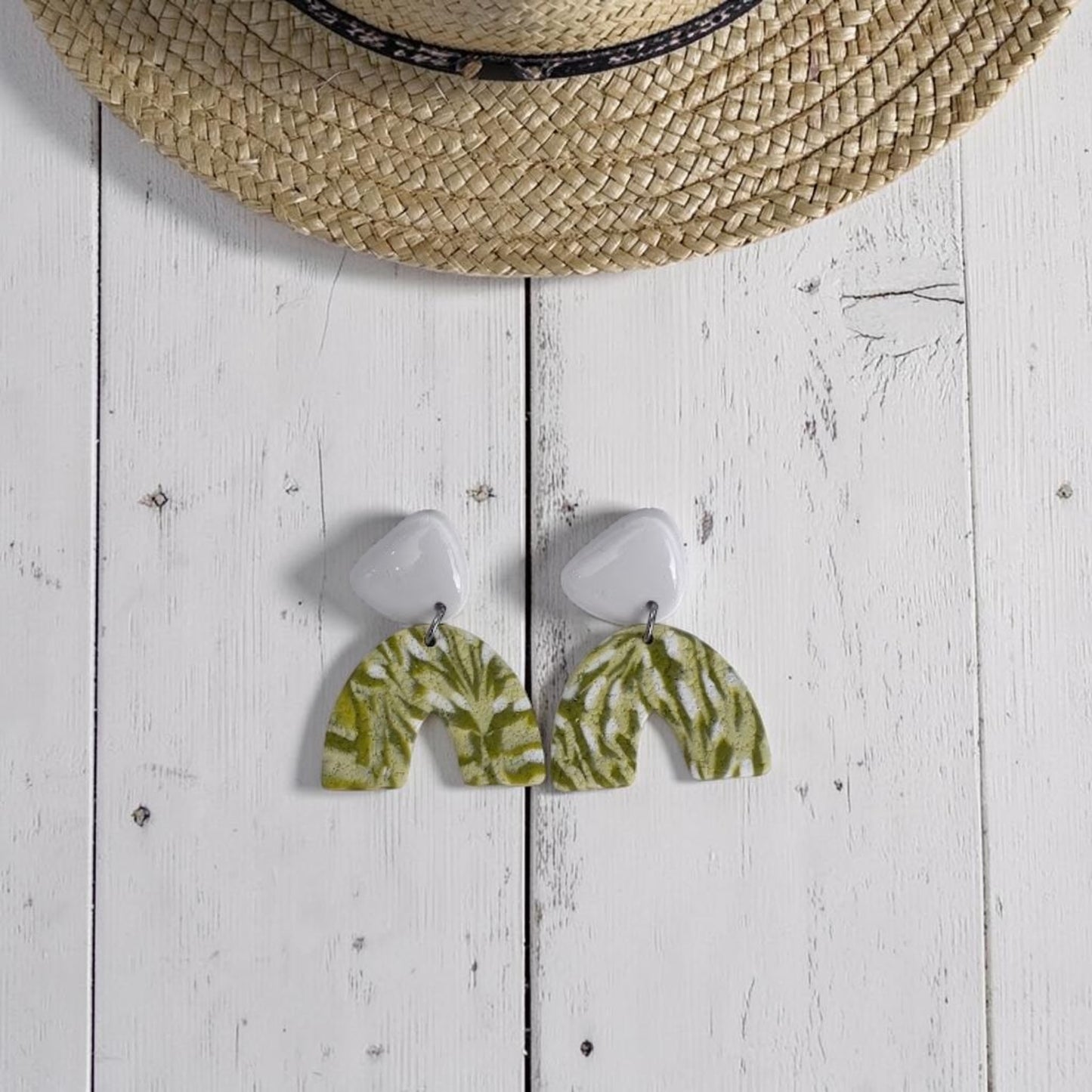 Pendientes artesanales de arcilla polimérica con diseño de arco con estampado marmoleado en verdes y blancos y pieza superior blanca acabada con resina UV, presentados sobre fondo de madera blanca con un detalle de un sombrero de rafia