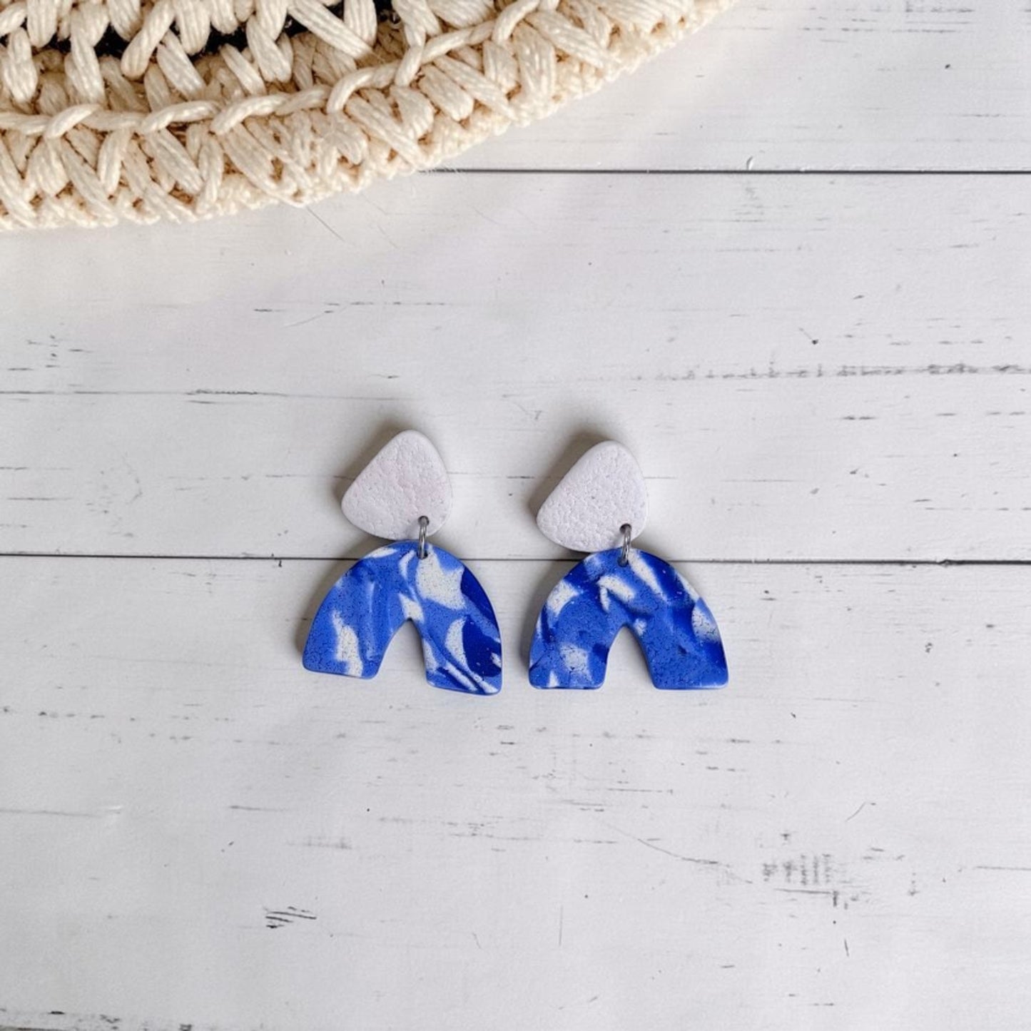 Pendientes artesanales de arcilla polimérica con diseño de arco con estampado en azules y blancos y pieza superior  con textura en blanco, presentados sobre fondo de madera blanca y detalle de sobrero de rafia.