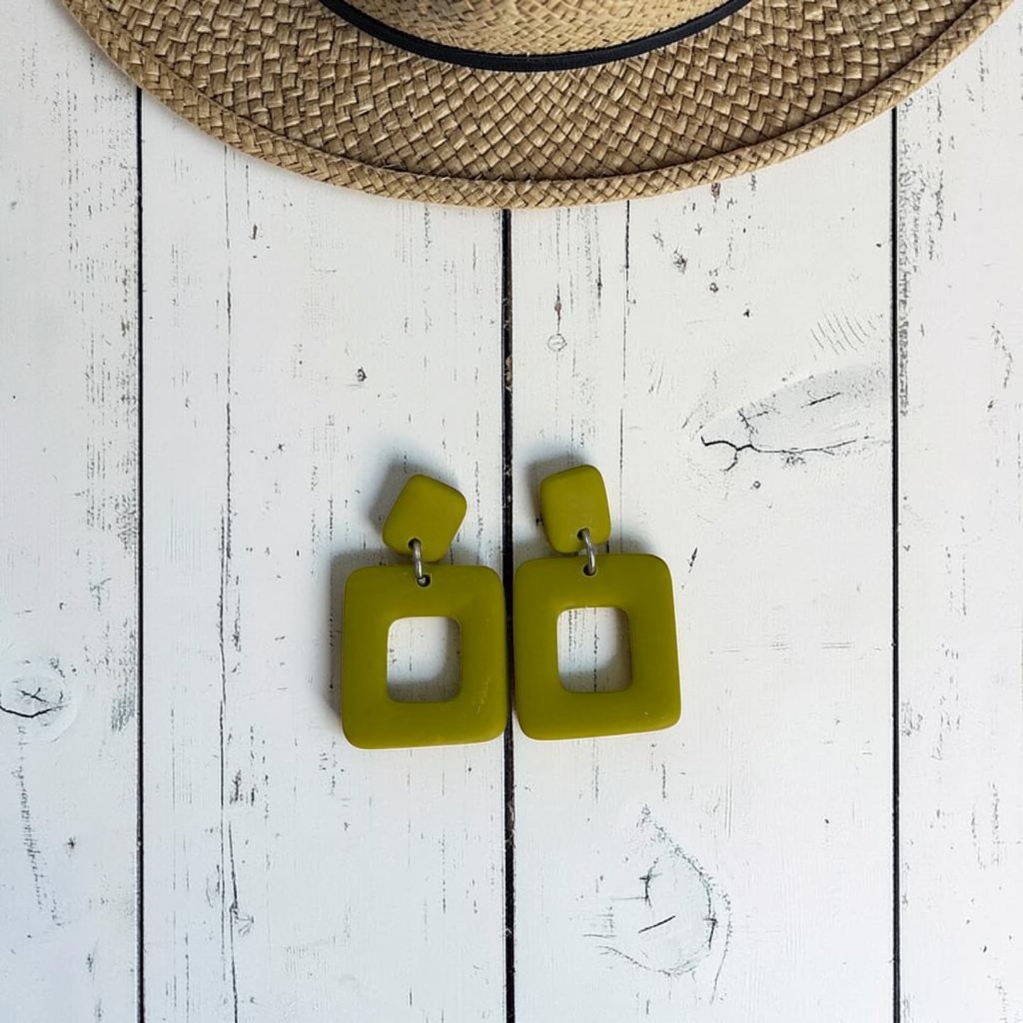 Pendientes Cubuk verde oliva de arcilla polimérica, vista del par completo sobre fondo de madera blanca con sombrero de paja