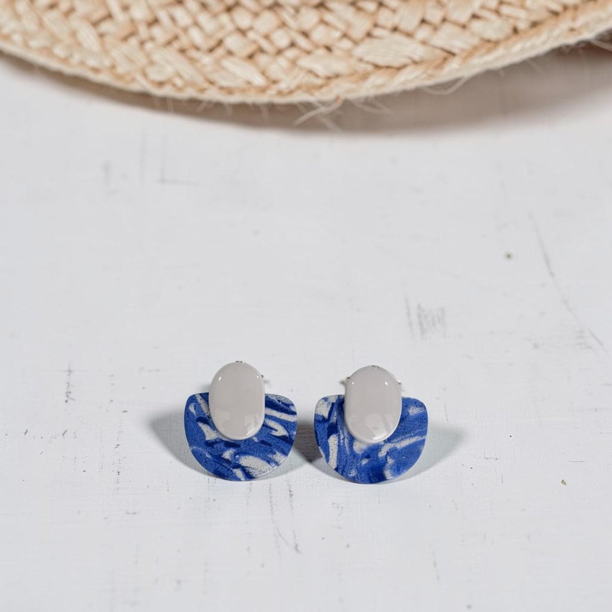 Pendientes Toffee azul y blanco de arcilla polimérica con efecto marmolado, vista del par completo sobre fondo blanco con sombrero de paja