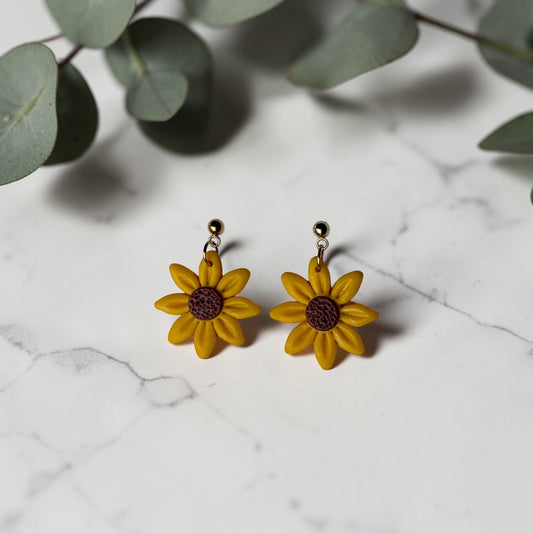 Pendientes artesanales de arcilla polimérica en forma de girasol amarillo con centro marrón sobre fondo de mármol blanco con hojas de eucalipto