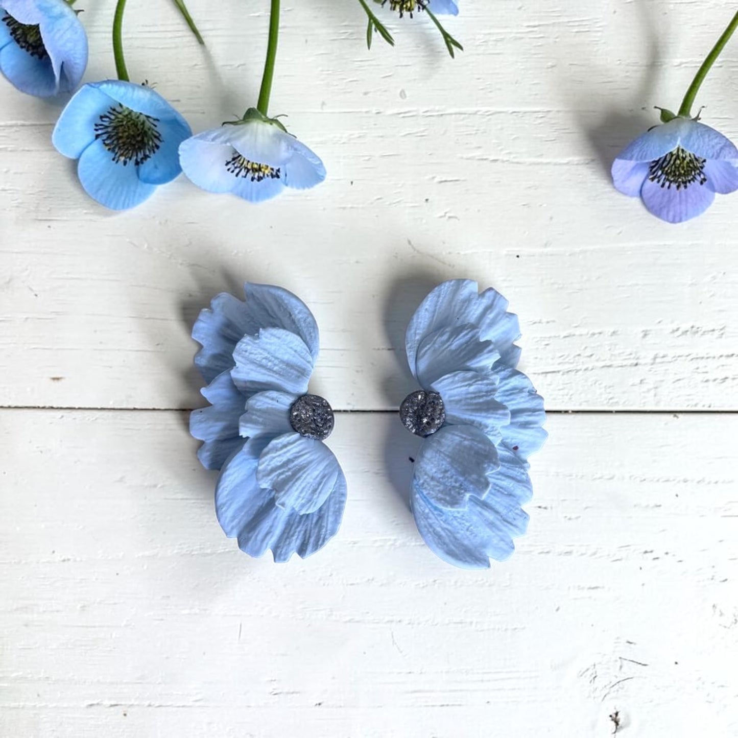 Pendientes artesanales Solleric de media flor azul nube en arcilla polimérica, sobre madera blanca con flores naturales