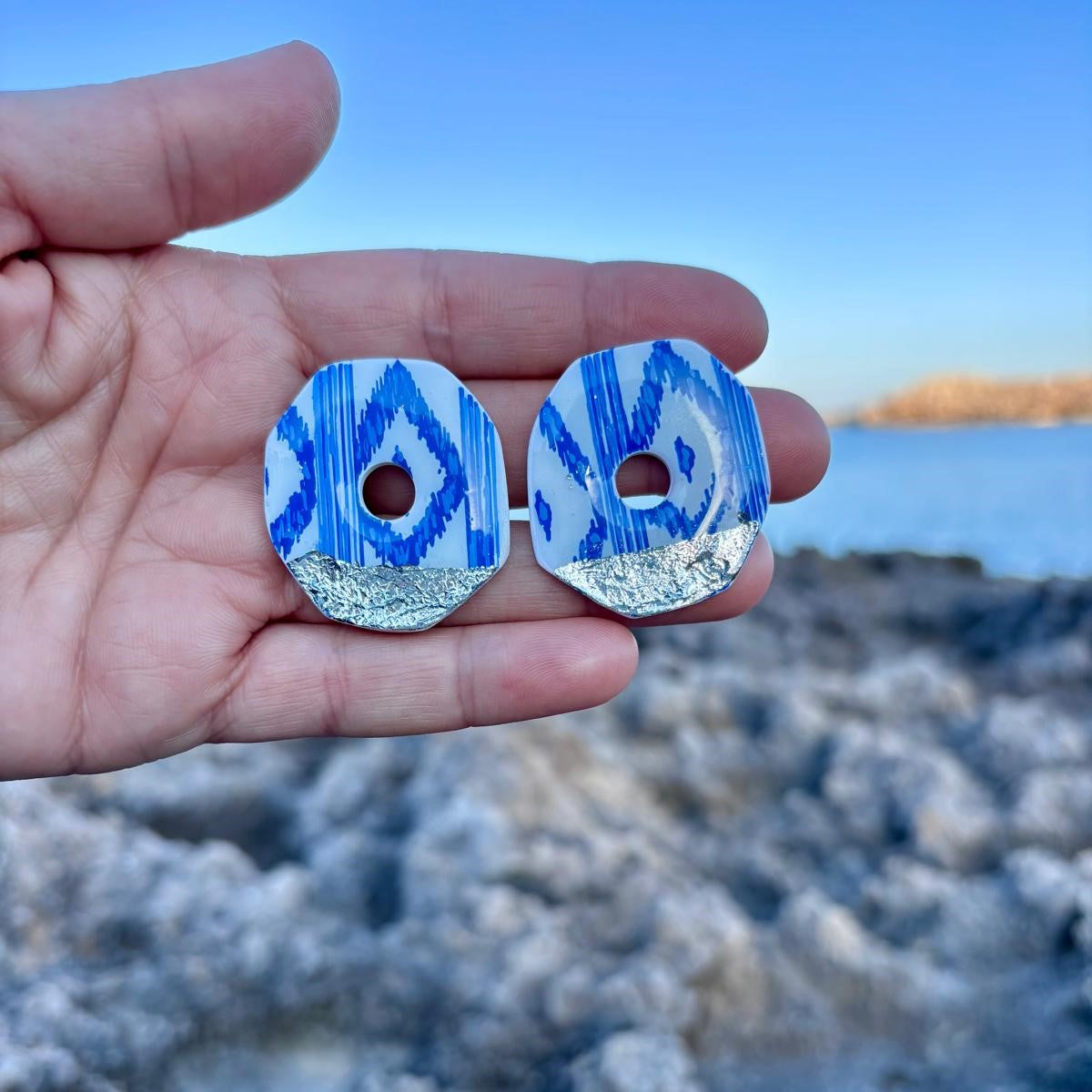 Primer plano de pendientes de botón artesanales con diseño de lenguas mallorquinas azules y base plateada, sostenidos en palma de la mano para mostrar su tamaño y acabado detallado frente al mar.
