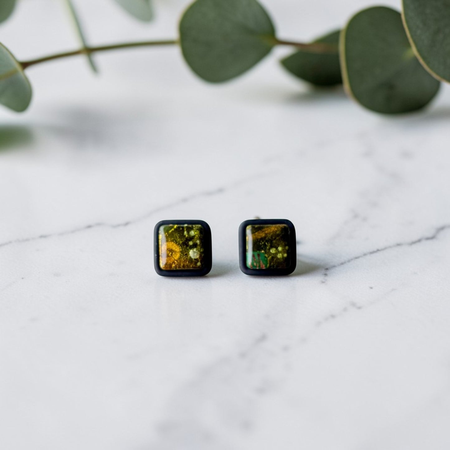 Pendientes artesanales tipo botón de arcilla polimérica acabado con resina, cuadrado verde, dorado y negro sobre fondo de mármol blanco con eucalipto