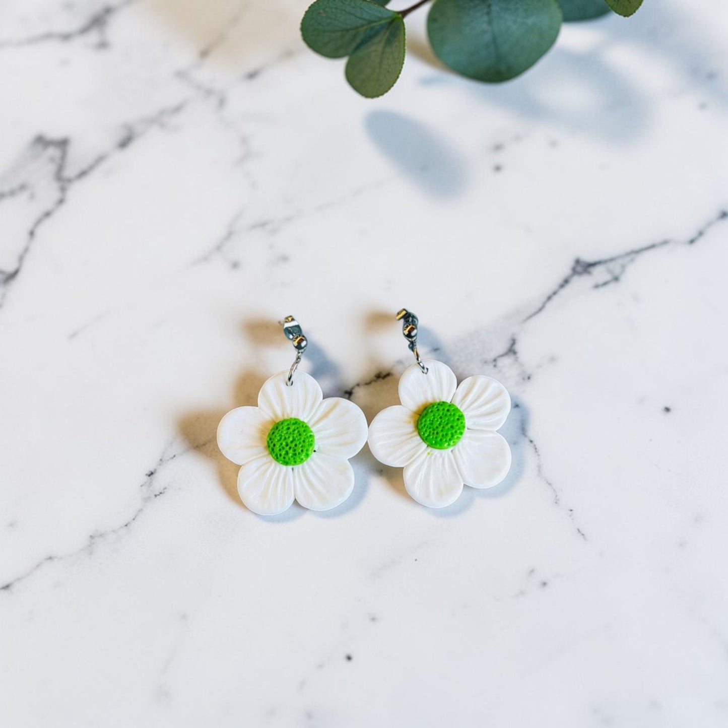 Pendientes artesanales de arcilla polimérica con forma de margarita blanca y centro verde sobre fondo de mármol blanco con eucalipto