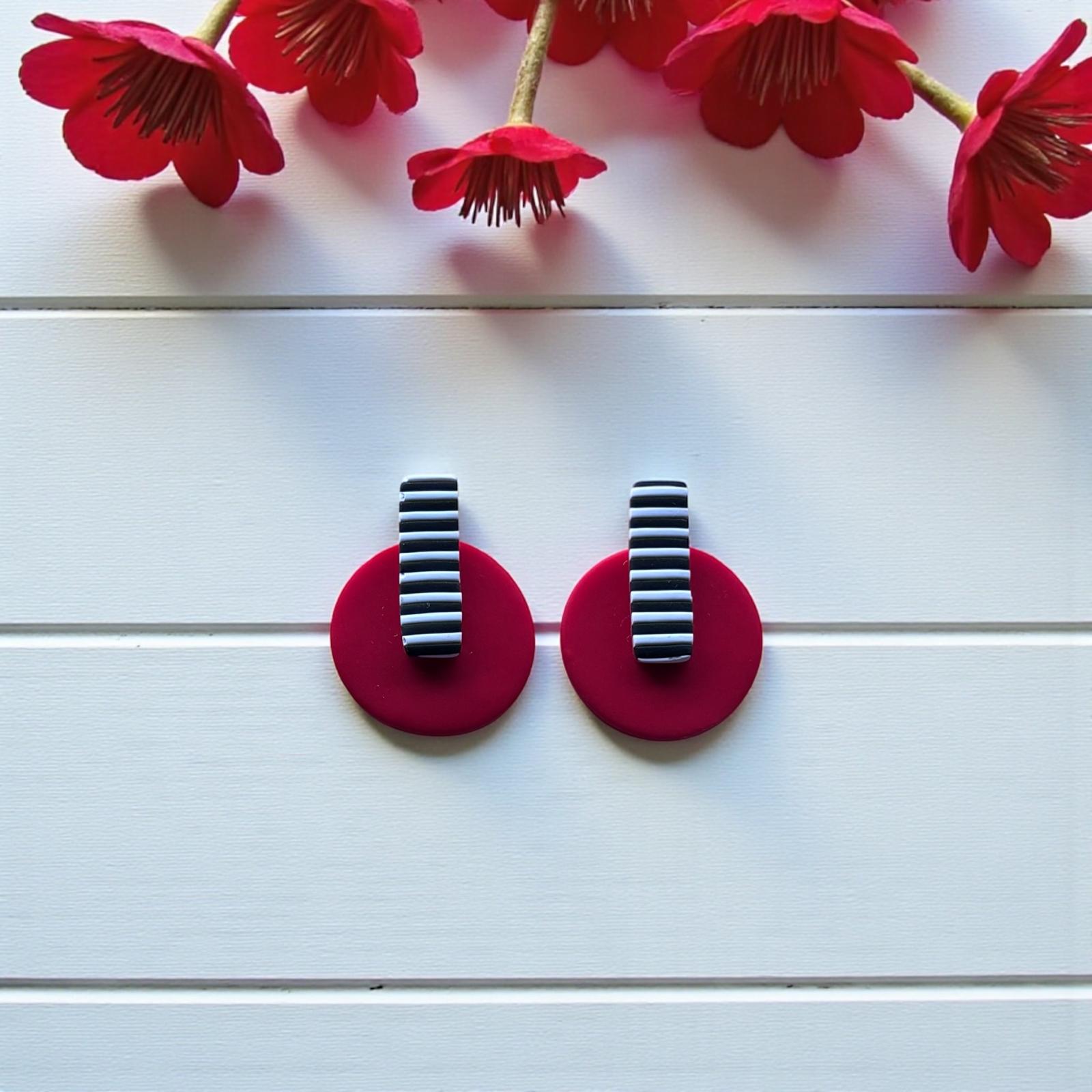 Primer plano de pendientes de diseño con base circular roja y detalle rectangular listado en blanco y negro sobre fondo de madera blanca con flores
