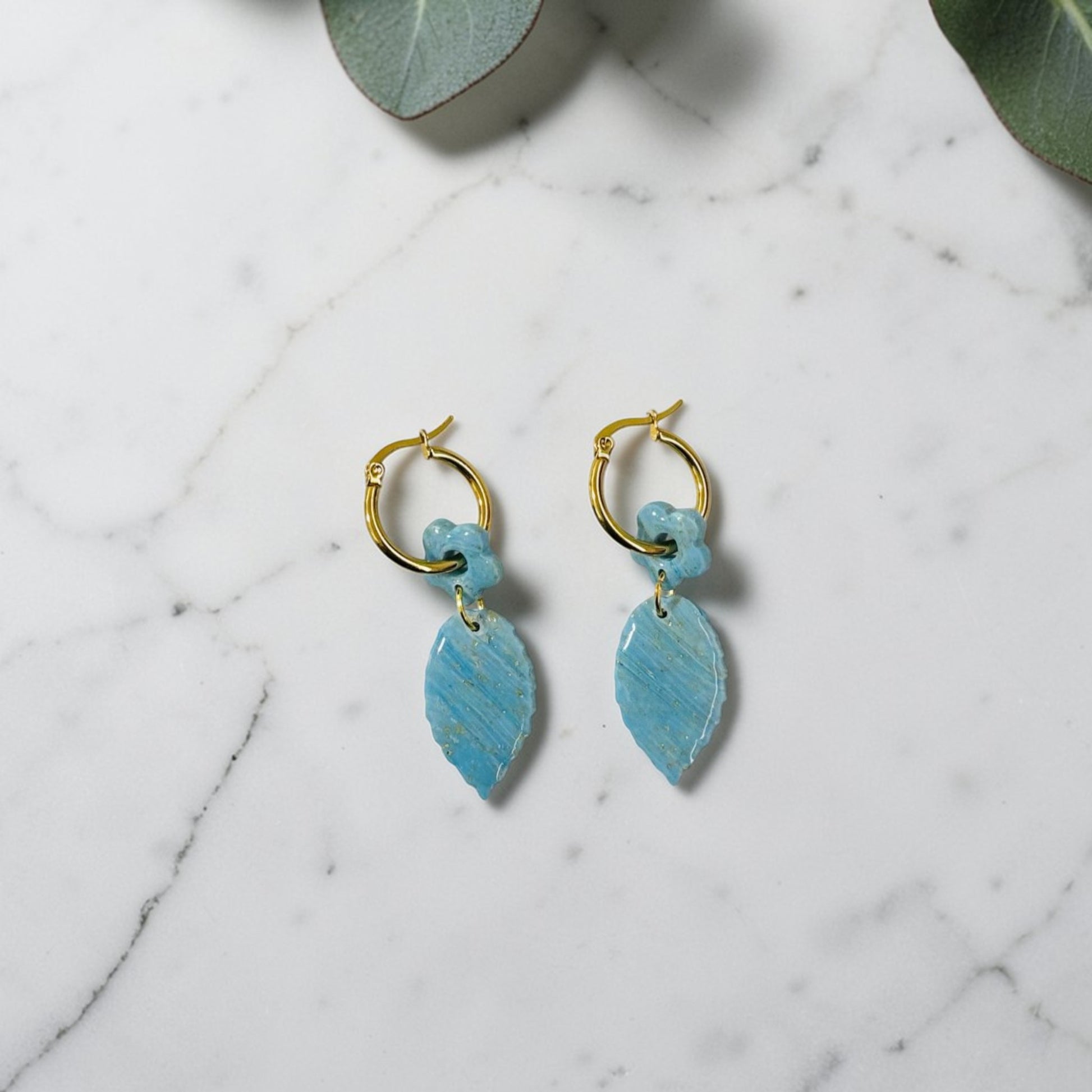Pendientes artesanales de arcilla polimérica con charm de hoja y flor azul turquesa y dorada sobre fondo de mármol blanco