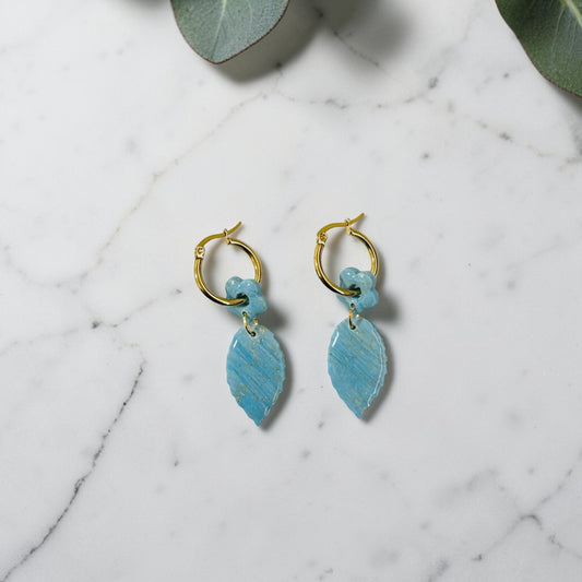 Pendientes artesanales de arcilla polimérica con charm de hoja y flor azul turquesa y dorada sobre fondo de mármol blanco