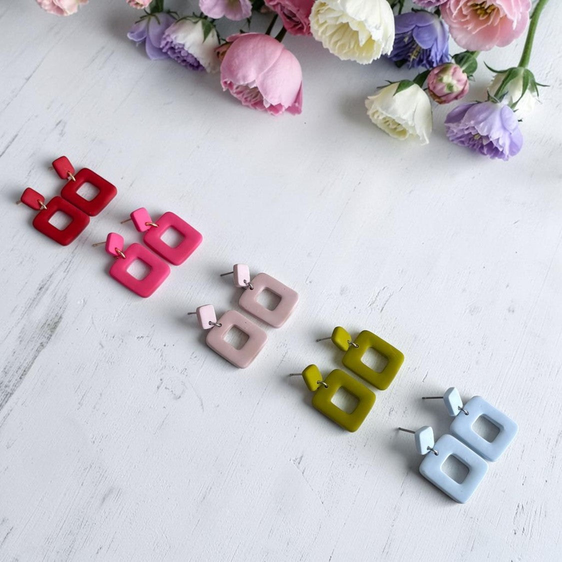 Pendientes Cubuk de arcilla polimérica en varios colores: rojo, rosa coral, beige, verde oliva y azul nube, sobre fondo blanco con flores