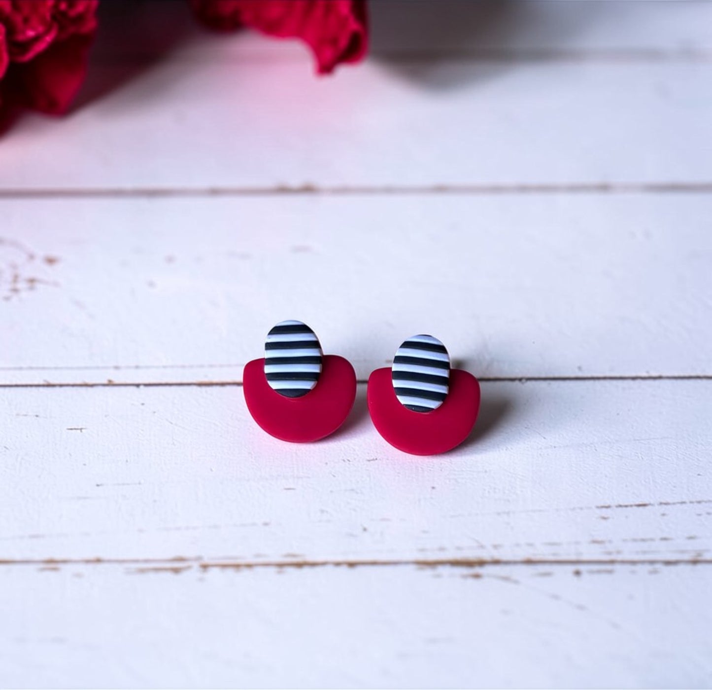 Pendientes artesanales de arcilla polimérica estilo geométrico con forma de medio círculo rojo y óvalo a rayas blancas y negras sobre fondo blanco de madera con flores rojas