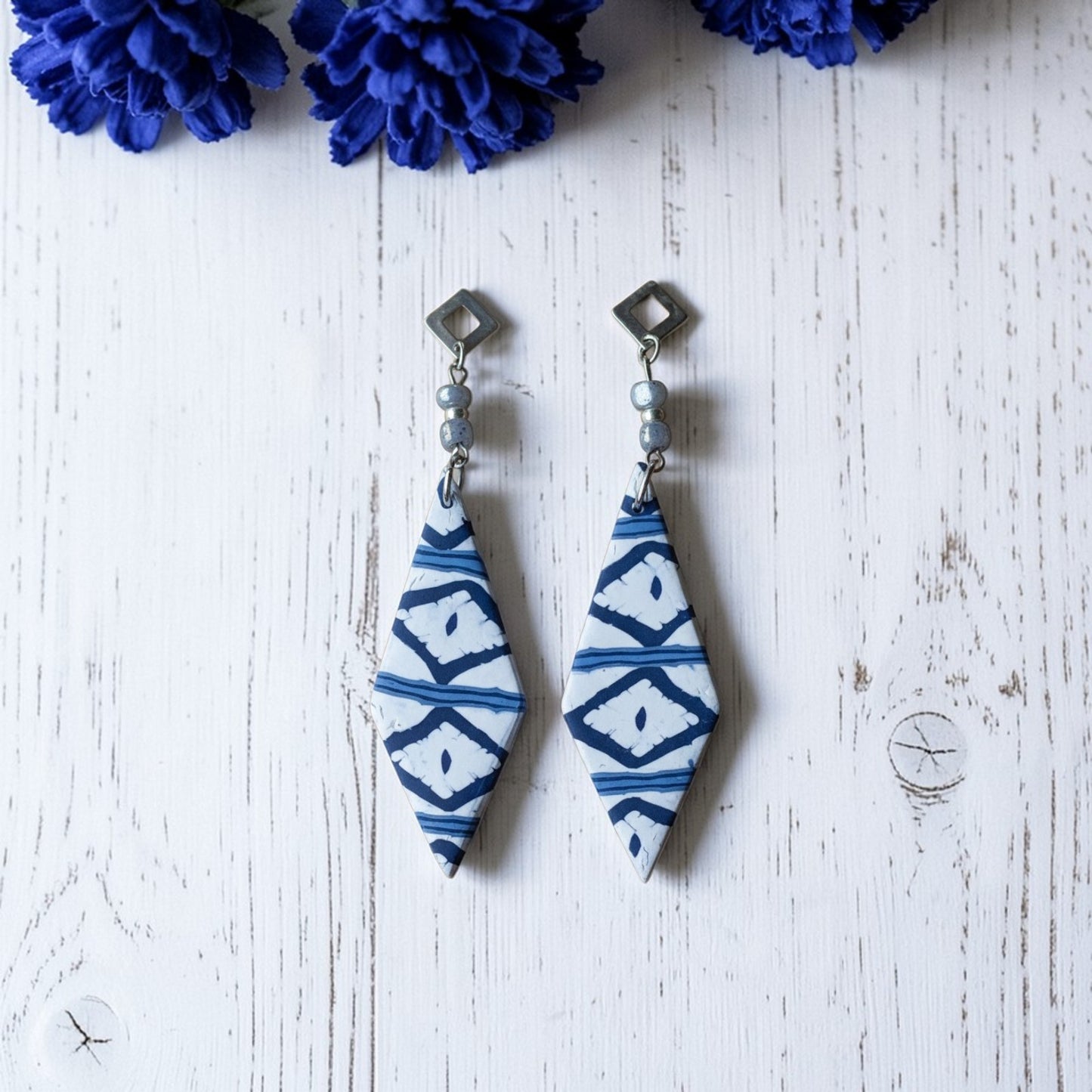 Pendientes largos artesanales Llengos Sal - Rombo largo de arcilla polimérica en azul marino y blanco con cuentas de vidrio celestes y fornitura bañada en plata, sobre fondo de madera blanca con flores azules. Colección Esencia Mallorquina de Pm_Art.
