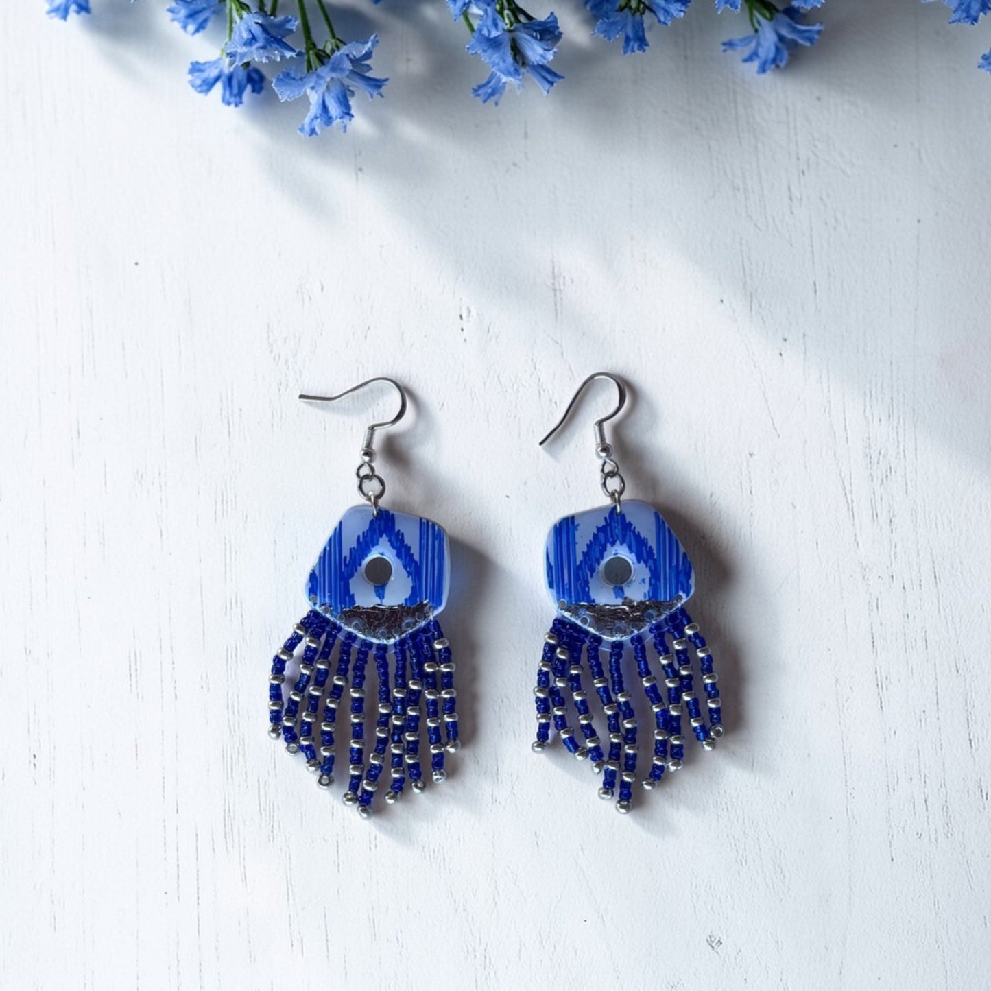 Pendientes largos artesanales con flecos de cuentas azules y diseño de lenguas mallorquinas sobre fondo de madera blanca con flores