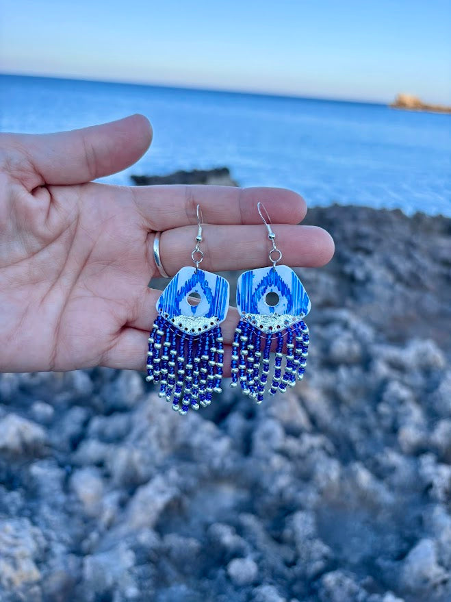 Primer plano de pendientes largos de la colección Esencia Mallorquina. Se detallan los flecos de cuentas azules y plateadas y la pieza superior de arcilla polimérica sostenidos en la palma de la mano.