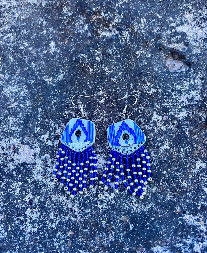 Pareja de pendientes largos de la colección Esencia Mallorquina sobre piedra natural. Destacan los flecos de cuentas azules y plateadas y el diseño artesanal inspirado en las lenguas mallorquinas tradicionales.