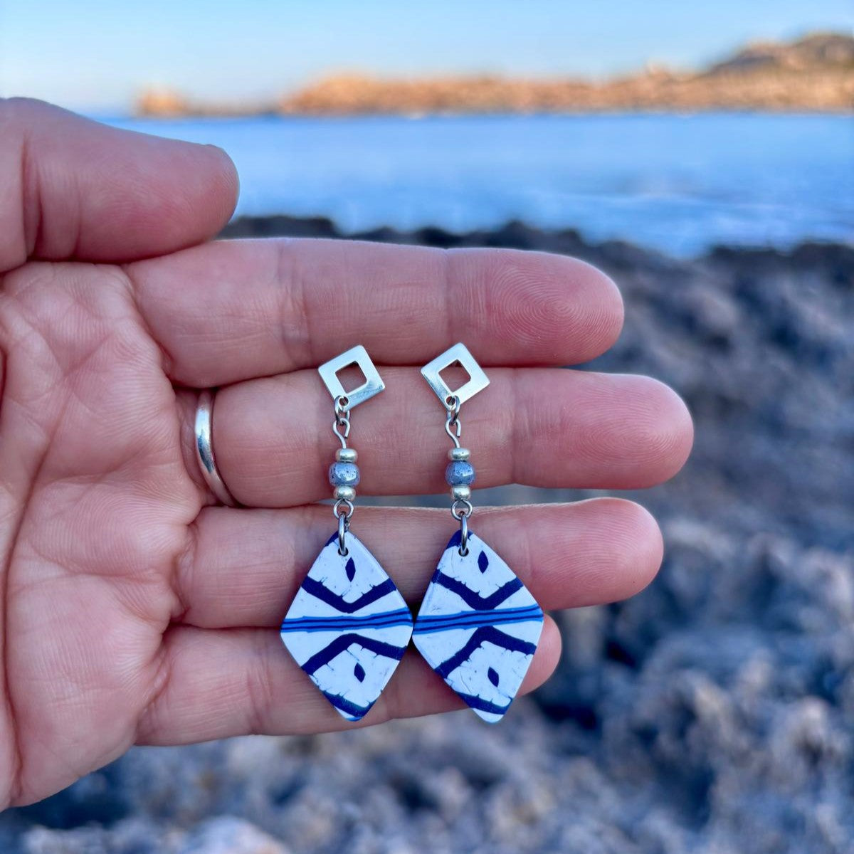 Pendientes cortos artesanales Llengos Sal - Rombo corto de arcilla polimérica en azul marino y blanco con cuentas de vidrio celestes y fornitura bañada en plata, sostenidos en mano con el mar Mediterráneo y la costa de Mallorca al fondo. Colección Esencia Mallorquina de Pm_Art.
