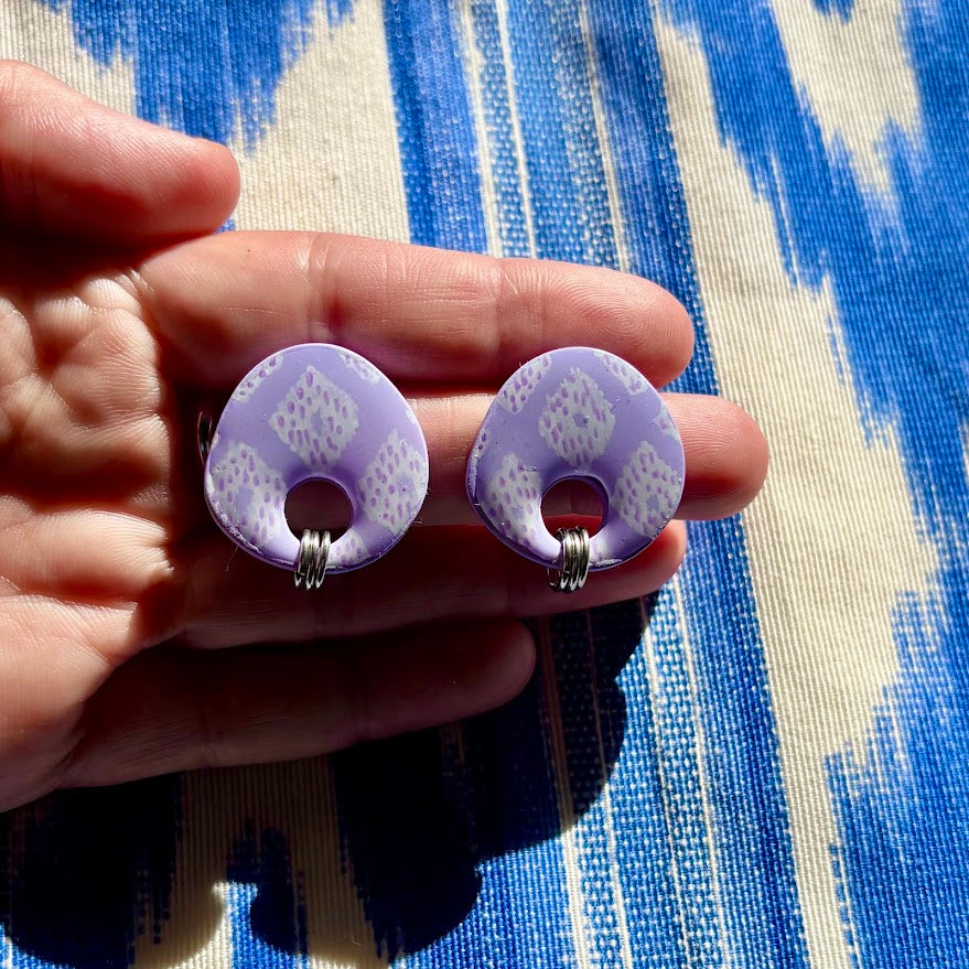 Pendientes llengos Cel color lila sostenidos en mano sobre tela de lenguas mallorquinas para apreciar la tradición plasmada en ellos