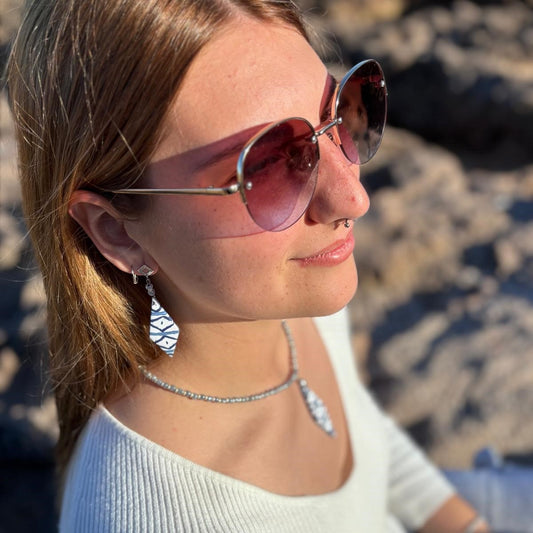 Modelo luciendo los pendientes largos artesanales Llengos Sal - Rombo largo de arcilla polimérica en azul y blanco con fornitura bañada en plata y collar de cuentas plateadas, con gafas de sol redondas y fondo de roca natural de Mallorca. Colección Esencia Mallorquina de Pm_Art.
