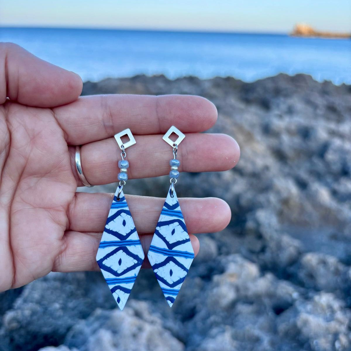 Pendientes largos artesanales Llengos Sal - Rombo largo de arcilla polimérica en azul marino y blanco con cuentas de vidrio celestes y fornitura bañada en plata, sostenidos en mano con la costa rocosa y el mar Mediterráneo de Mallorca al fondo. Colección Esencia Mallorquina de Pm_Art.
