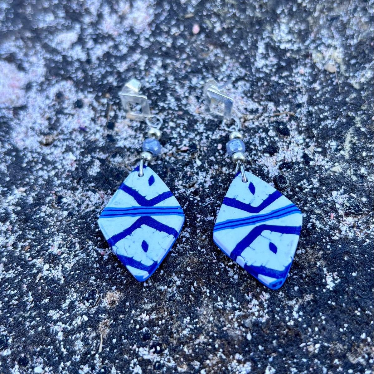 Pendientes cortos artesanales Llengos Sal - Rombo corto de arcilla polimérica en azul marino y blanco con cuentas de vidrio celestes y fornitura bañada en plata, sobre roca oscura de Mallorca. Colección Esencia Mallorquina de Pm_Art.
