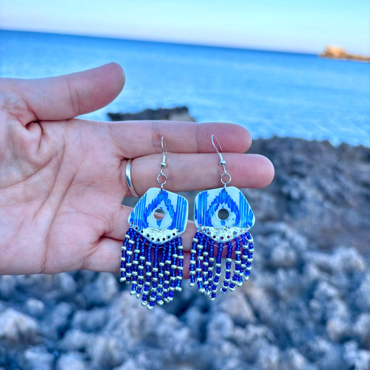 Primer plano de pendientes largos artesanales con flecos de cuentas azules y plateadas, sostenidos en la ano frente a un paisaje costeo. El diseño superior muestra el estampado de lenguas mallorquinas de la colección Esencia Mallorquina.
