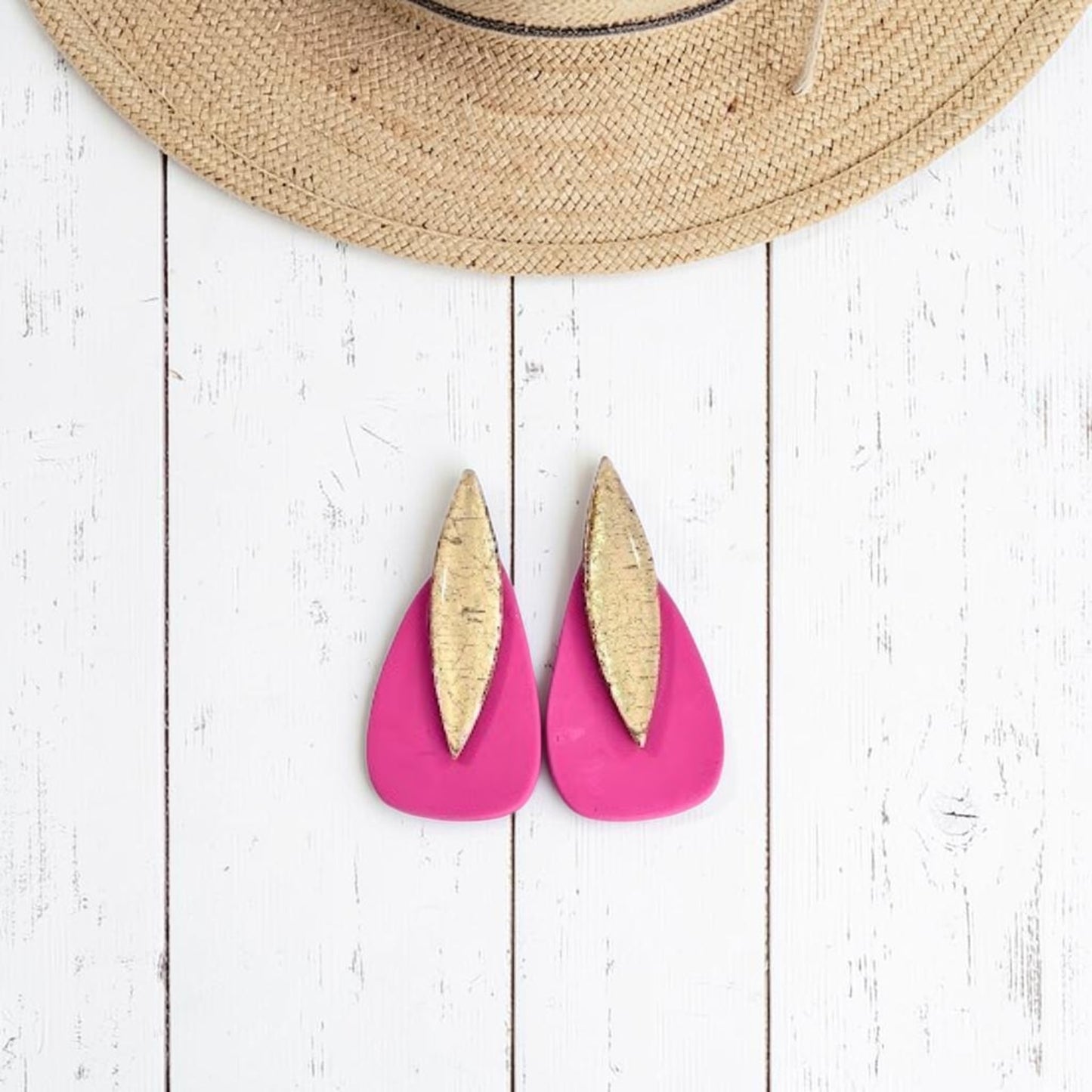 Pendientes modelo Sail color rosa coral de arcilla polimérica, diseño gota bicolor artesanal combinado con dorado, sobre fondo de madera y sombrero de paja en la parte superior de la imagen