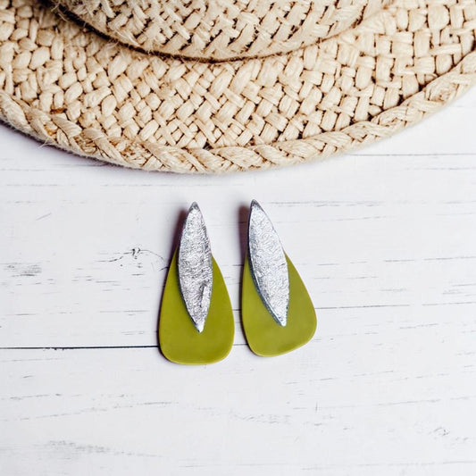 Pendientes Sail bicolor en verde oliva y plata, arcilla polimérica artesanal, forma de gota con textura mate en la base, sobre fondo de madera blanca y sombrero de paja en la parte superior de la imagen