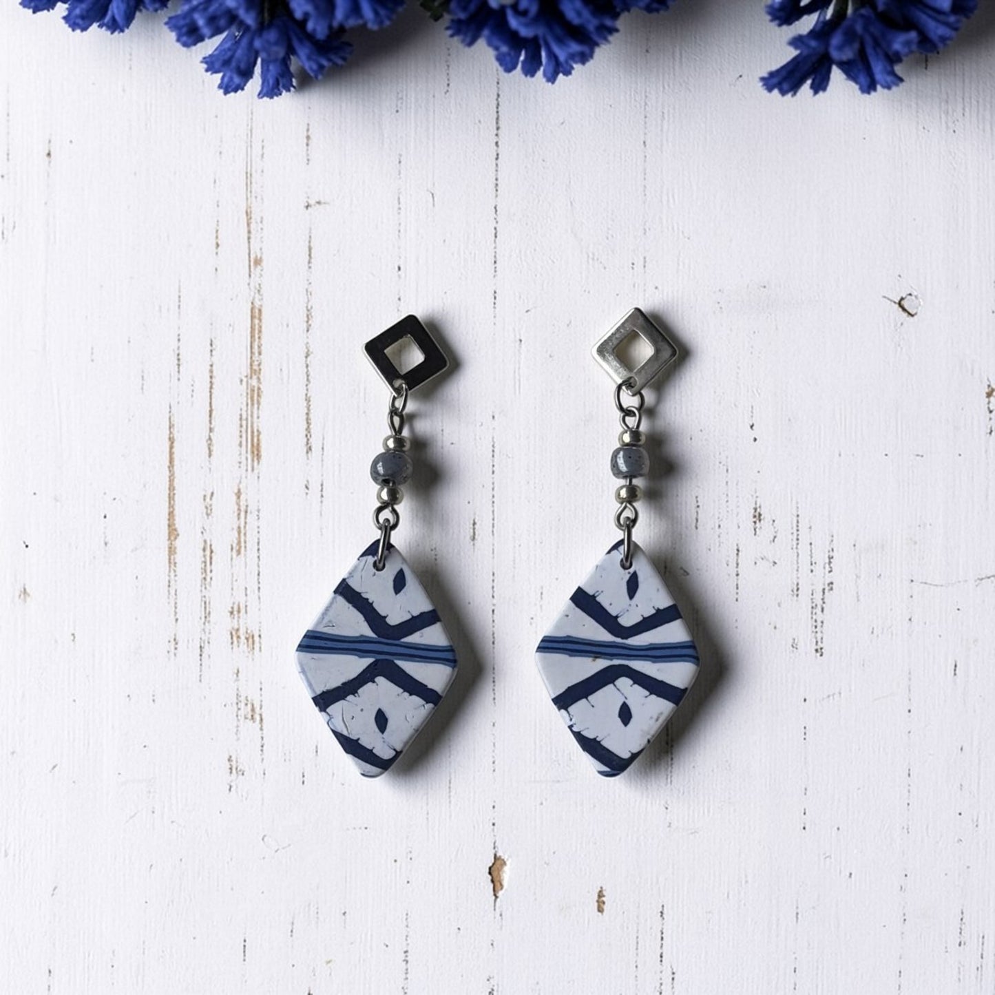 Pendientes cortos artesanales Llengos Sal - Rombo corto de arcilla polimérica en azul marino y blanco con cuentas de vidrio celestes y fornitura bañada en plata, sobre fondo de madera blanca con flores azules. Colección Esencia Mallorquina de Pm_Art.
