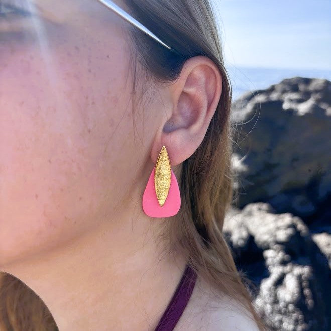 Modelo con pendientes artesanales Sail color rosa coral, detalle en oreja, fondo de rocas y mar